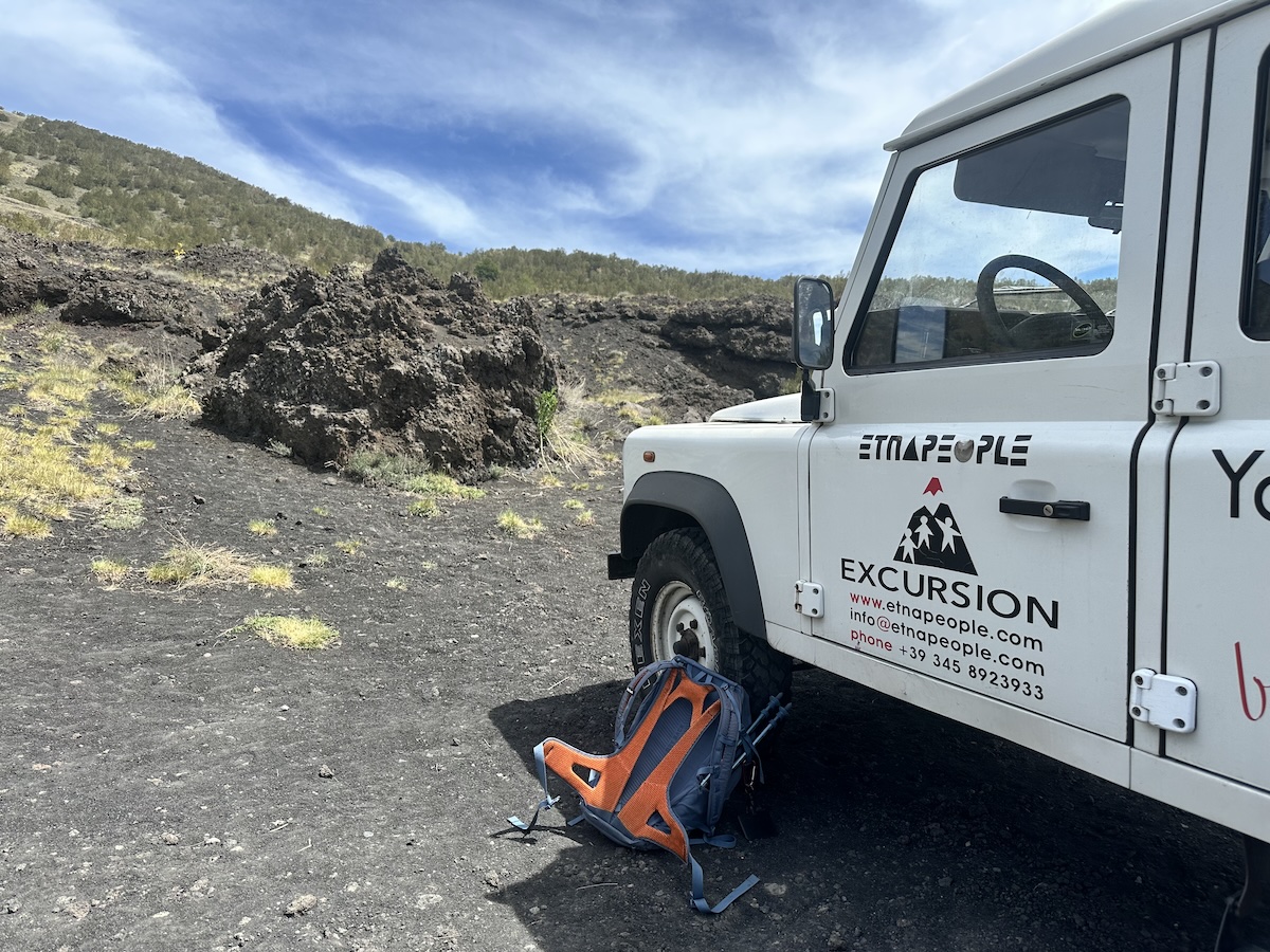 Am Ätna-Ausflug in Catania steht ein weißes EtnaPeople-Excursion Fahrzeug auf schwarzem Lavafeld, daneben ein orangefarbener Rucksack unter blauem Himmel.