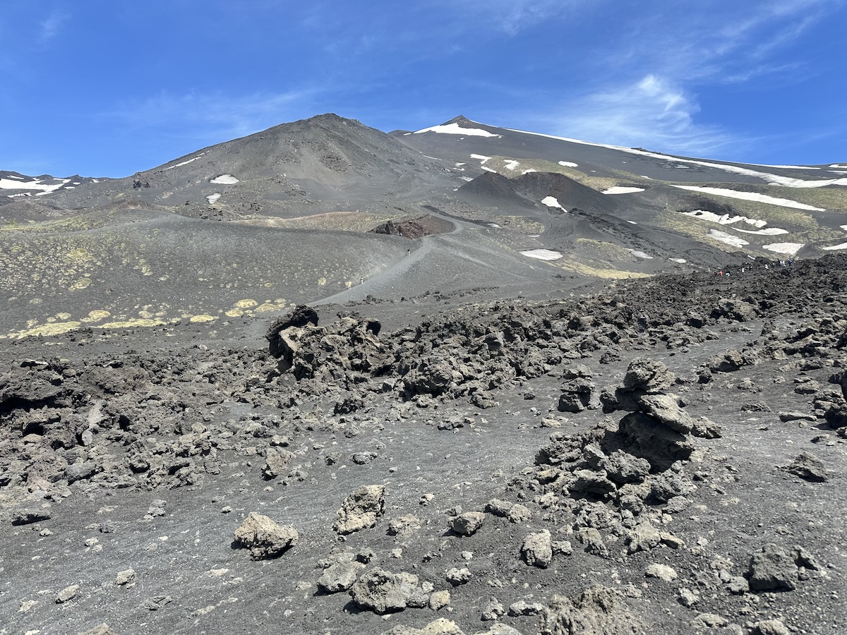 Auf dem Ätna, steinige grau-schwarze Vulkanlandschaft mit dunklen Lavasteinen und einigen Schneeflecken, darunter ein weites Kraterfeld. Tagesausflüge ab Catania