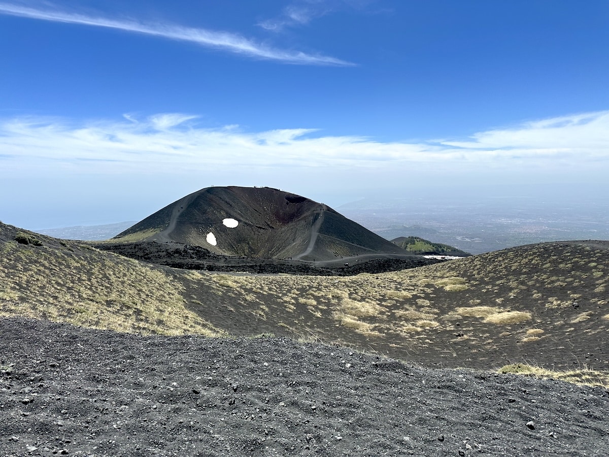 Blick auf den Ätna-Vulkan mit dunklem, grauem Lavagestein, zwei hellen Dampflöchern im Krater und weiter Sicht in Richtung Catania unter blauem Himmel.