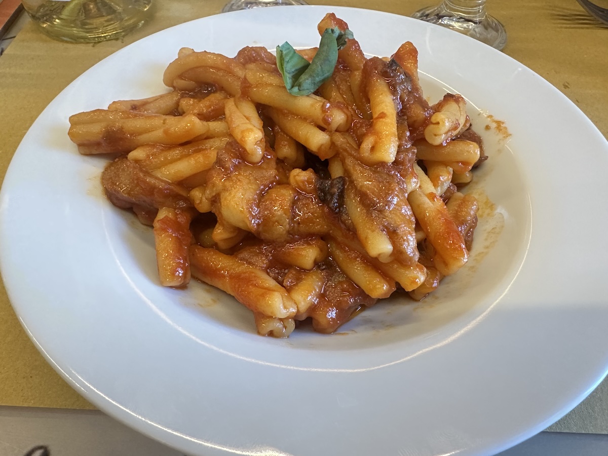 Auf einem weißen Teller liegen Pasta alla Norma mit tomatig-orangefarbener Sauce und bräunlichen, kräftig überbackenen Stücken, serviert in Catania.