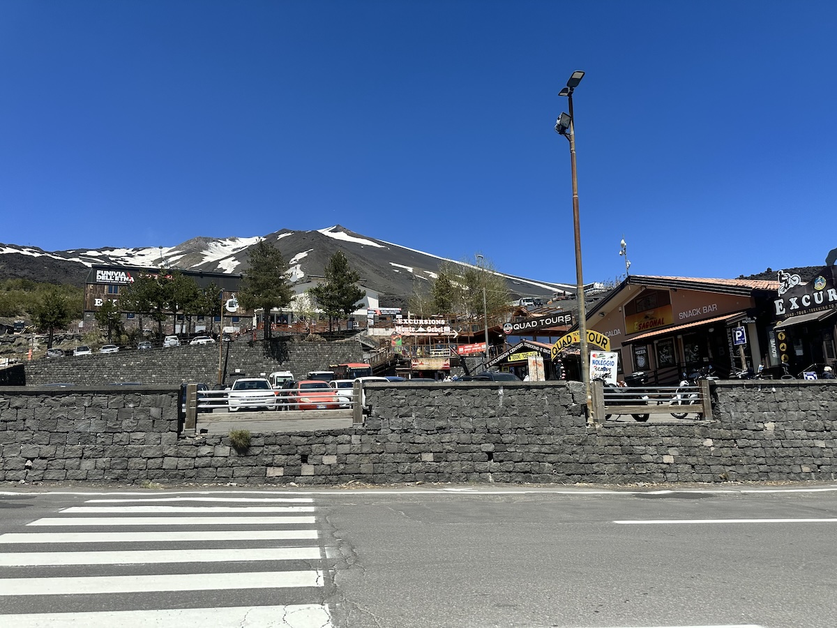 Vor der Kulisse des schneebedeckten Ätna steigen Gebäude und Schilder wie „Funivia dell’Etna“ und „Quad Tours“ in den blauen Himmel hinein.