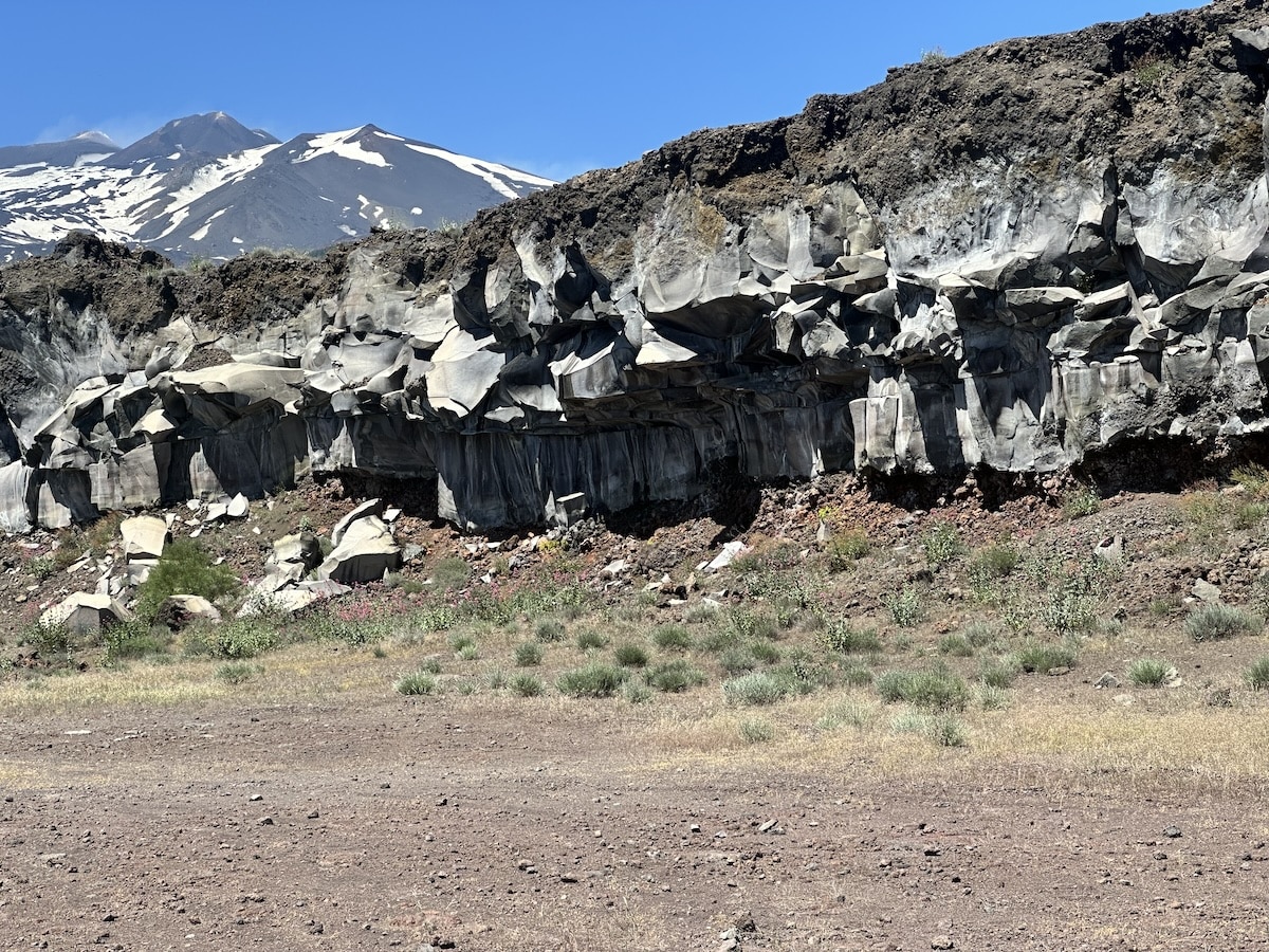 Im Vordergrund schwarze und grau-weiße Felswände einer Lavahöhle, im Hintergrund der Ätna mit schneebedecktem Gipfel vor blauem Himmel bei Catania