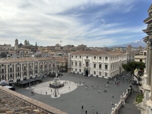 Blick über Catania auf den Piazza del Duomo im Centro Storico, mit Kuppeln über den Altstadt-Gebäuden, wolkigem Himmel und hellem Tageslicht.