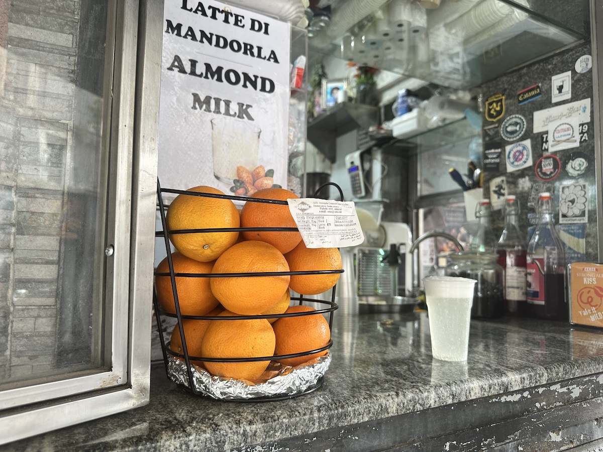 In der kühlen Theke von Catania stehen orangefarbene Orangen im Gestell, während „Latte di Mandorla / Almond Milk“ an der Front ausgeschildert ist.