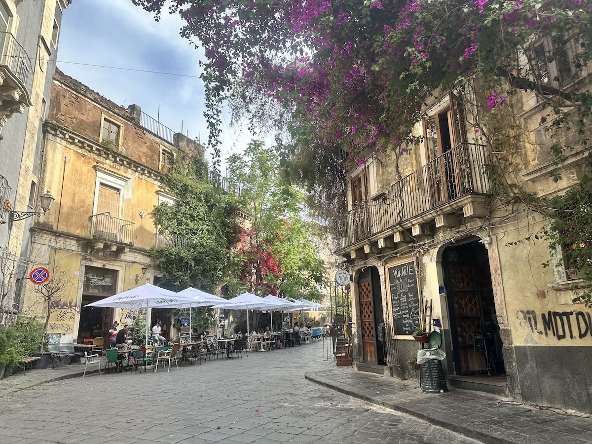 Zwischen gelblichen Fassaden in Catania und rund um den Piazza del Duomo lädt ein gepflasterter Platz mit Café-Tischen unter blühendem Lila zum Verweilen ein.