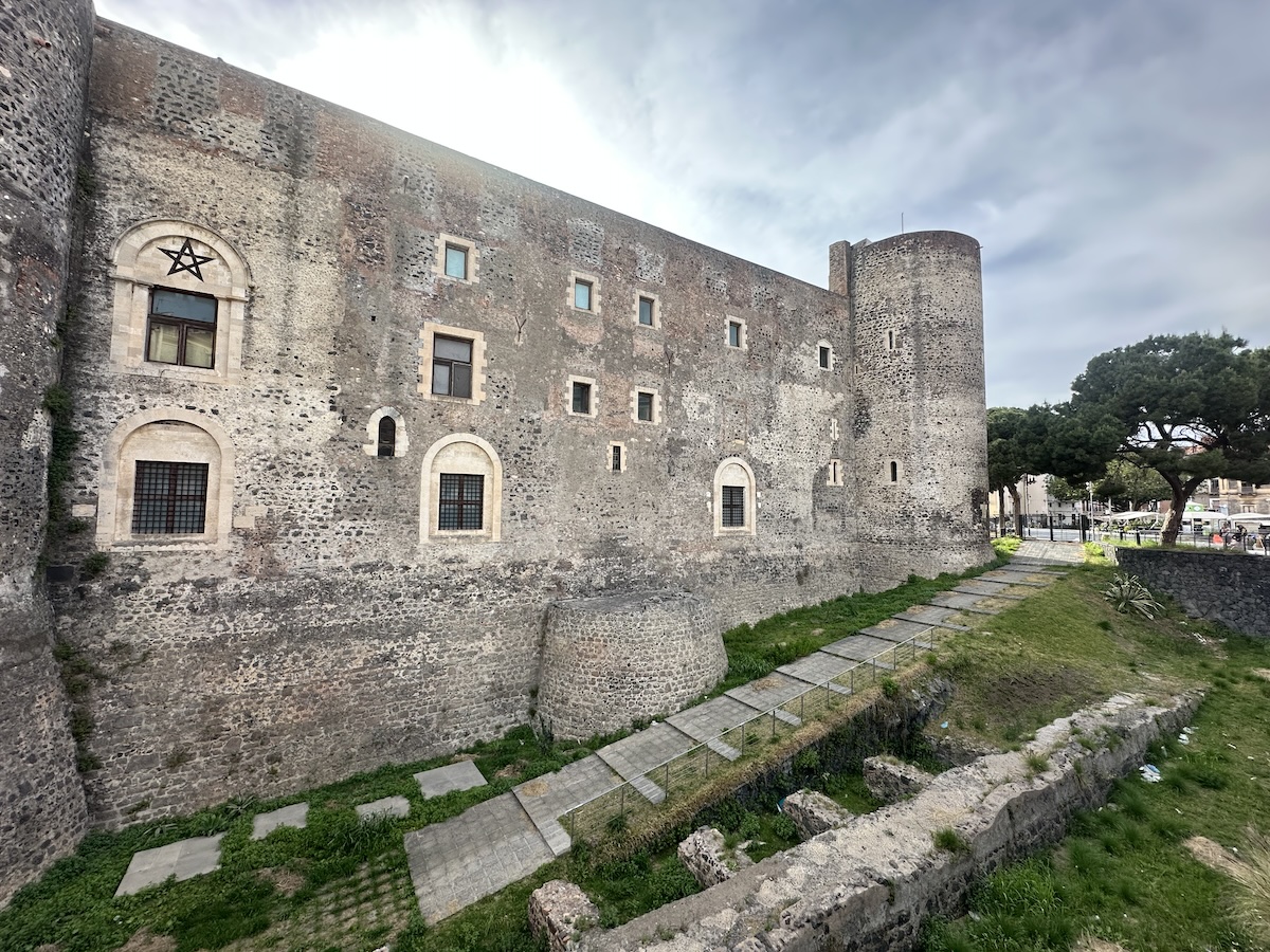 Castello Ursino mit dunklem Lavagestein, grauer Burgmauer und Rundturm, dazu schmale Fensteröffnungen und ein Sternsymbol an der Fassade unter Wolken.