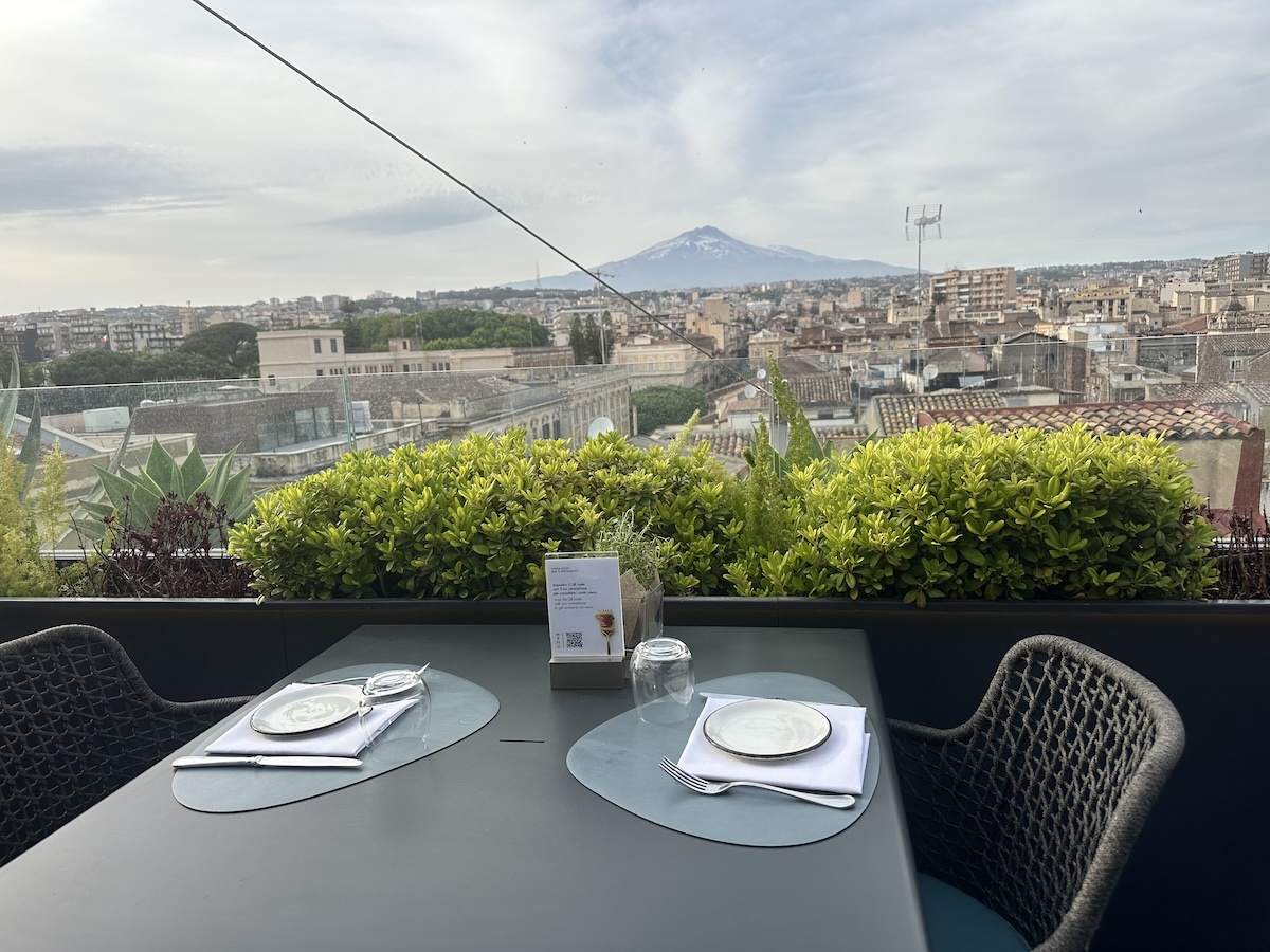 Auf einer Terrasse in Catania mit grünen Pflanzen stehen gedeckte Tische, während sich im Hintergrund der schneebedeckte Ätna unter bewölktem Himmel abzeichnet.
