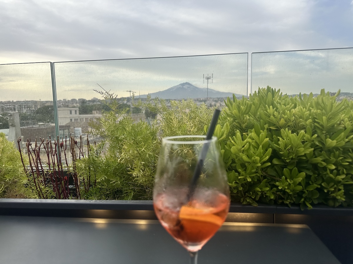 Orange Drink im Glas vor grünen Pflanzen am Fenster, mit Blick über Catania und Richtung Via Etnea bei wolkenverhangenem Himmel, ruhig und urban