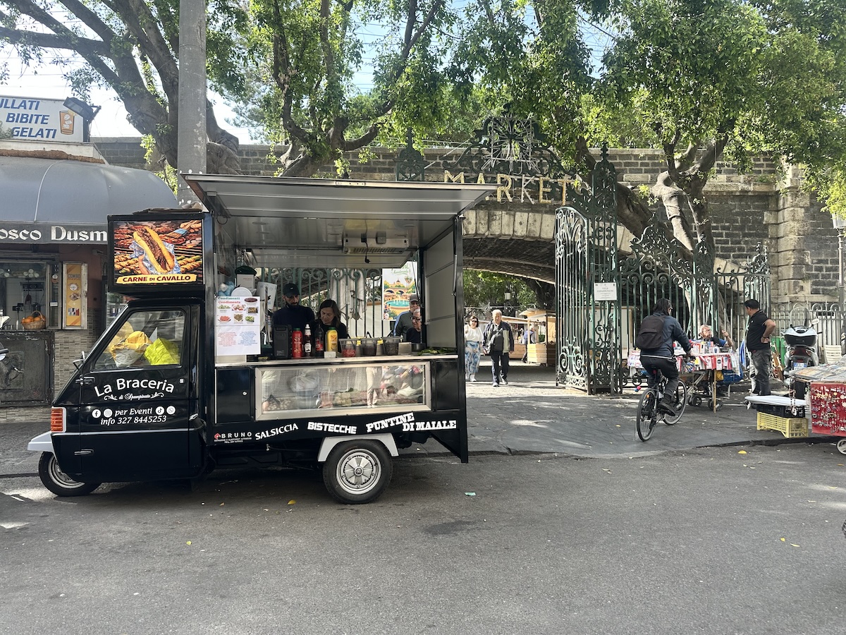 Vor Ort in Catania sorgt der Streetfood-Wagen „La Braceria“ mit schwarzem Aufbau und geöffnetem Grillbereich für Stimmung am Fischmarkt La Pescheria.