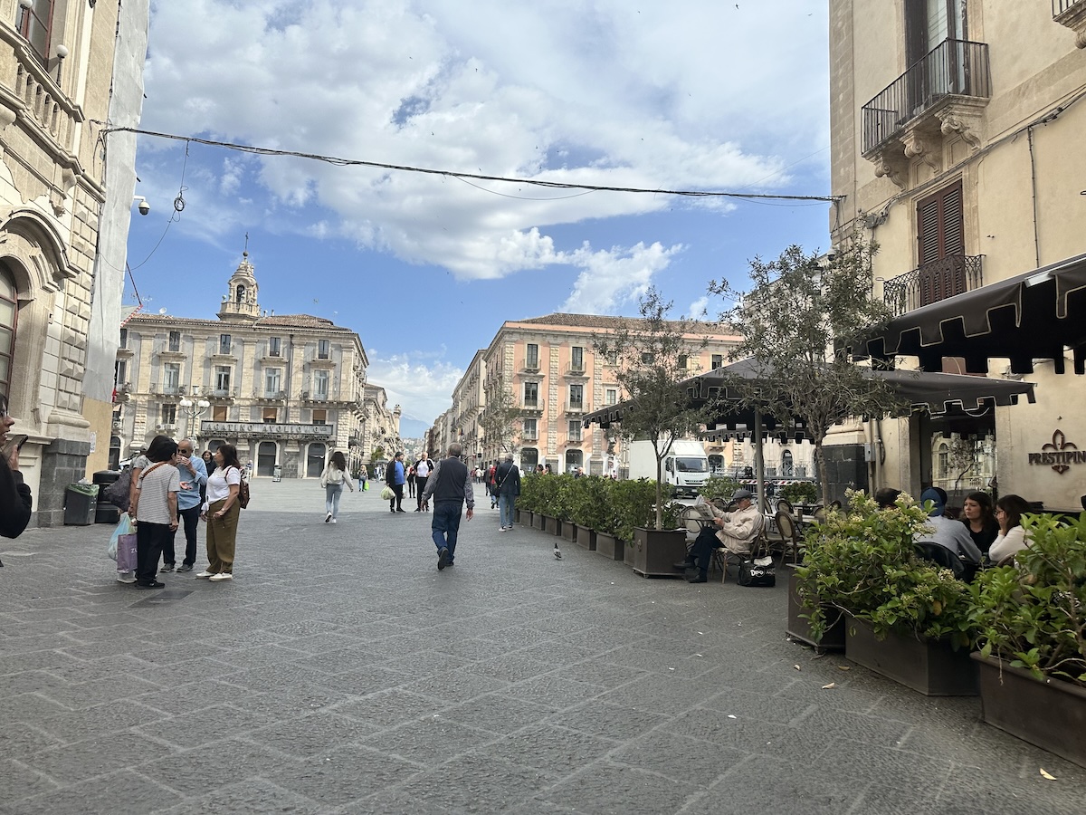 Zwischen historischen Gassen in Catania führt ein Weg aus Pflastersteinen an heller Steinfassade und Gebäuden vorbei, unter blauem Himmel mit Wolken.