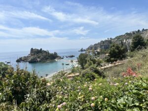 Blick von den Hügeln bei Taormina auf türkisblaues Meer und Isola Bella, mit Küste, Buchten und grünen Gärten unter blauem Himmel.