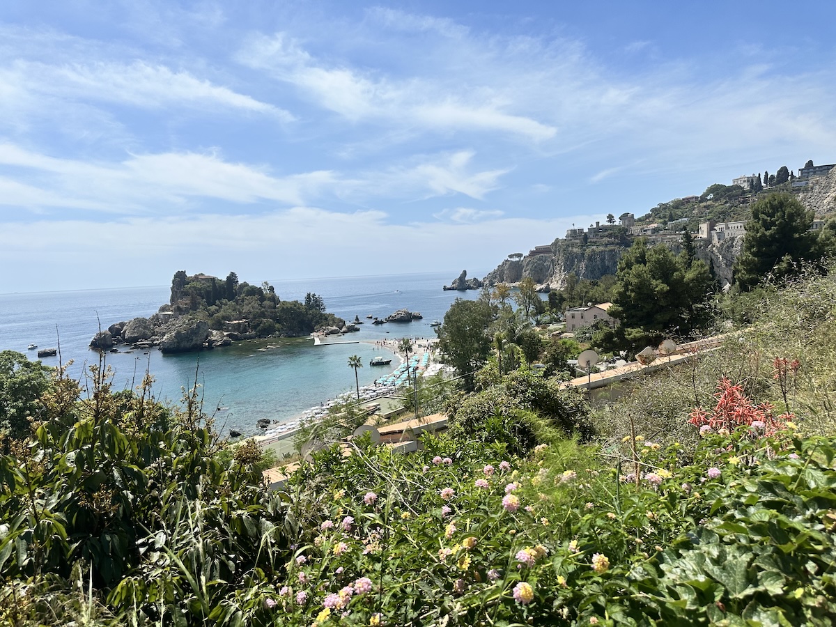 Blick von den Hügeln bei Taormina auf türkisblaues Meer und Isola Bella, mit Küste, Buchten und grünen Gärten unter blauem Himmel.
