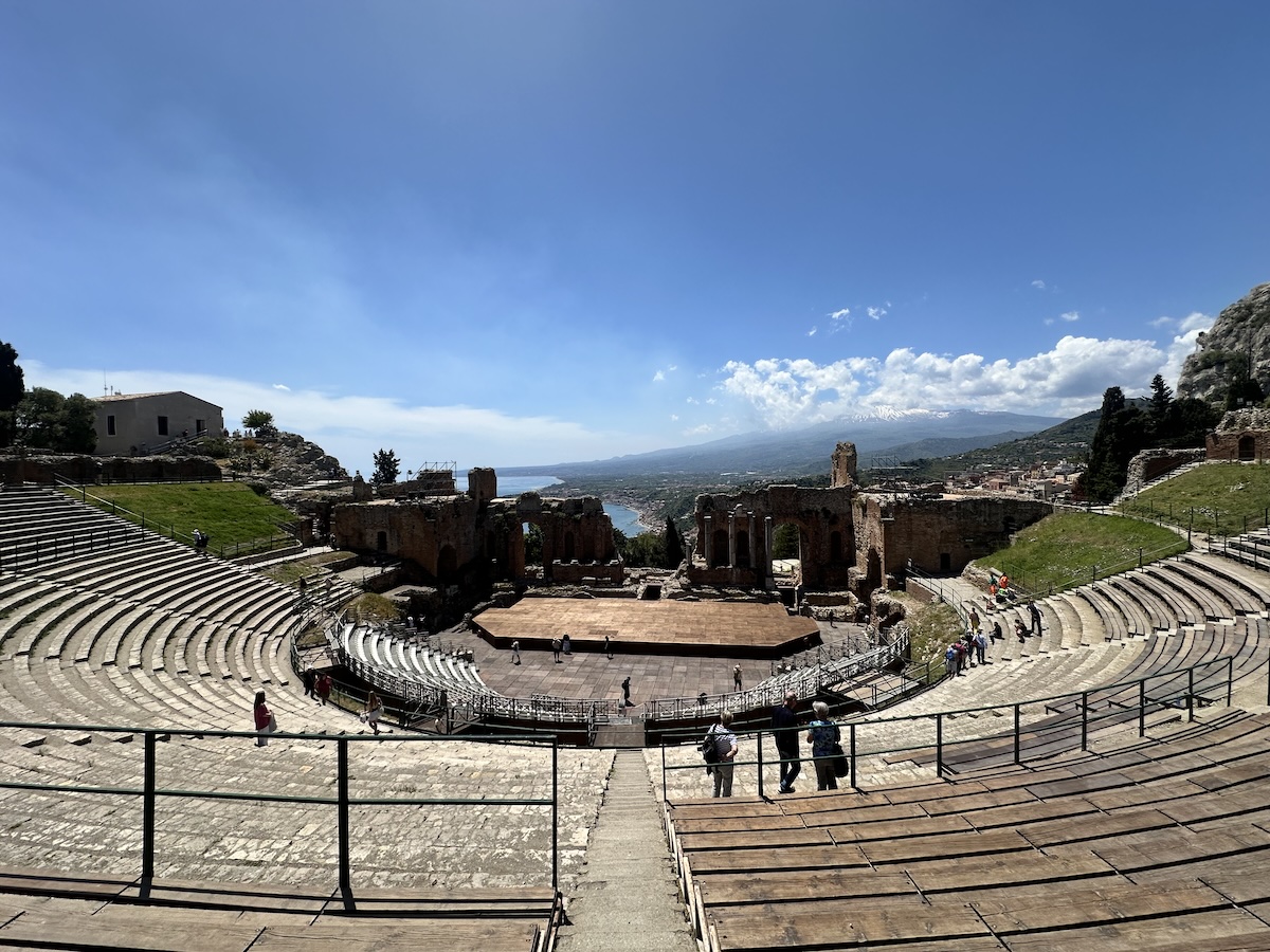 Blick über das steinerne Theater in Taormina mit Ruinen, grünen Hängen und weitem Meerblick, im Hintergrund der Ätna unter blauem Himmel.