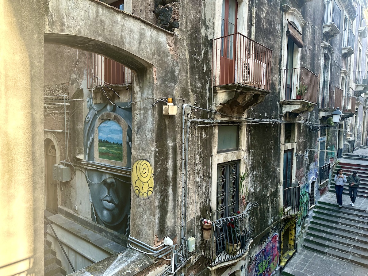 In Catania zeigt sich eine verwitterte, enge Gasse mit Graffiti an Mauern und Treppen, mit Balkonen und heruntergekommen wirkenden Fassaden im Altstadt-Charakter.