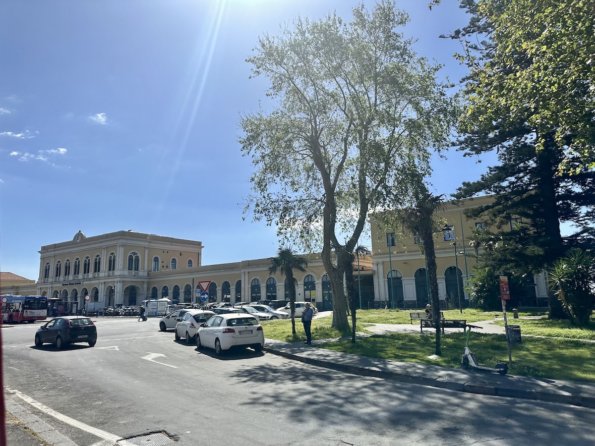 Vor einer großen, hellen Fassade in Catania stehen Autos und ruhige Bäume, während ein blauer Himmel den Bahnhofseingang umrahmt.