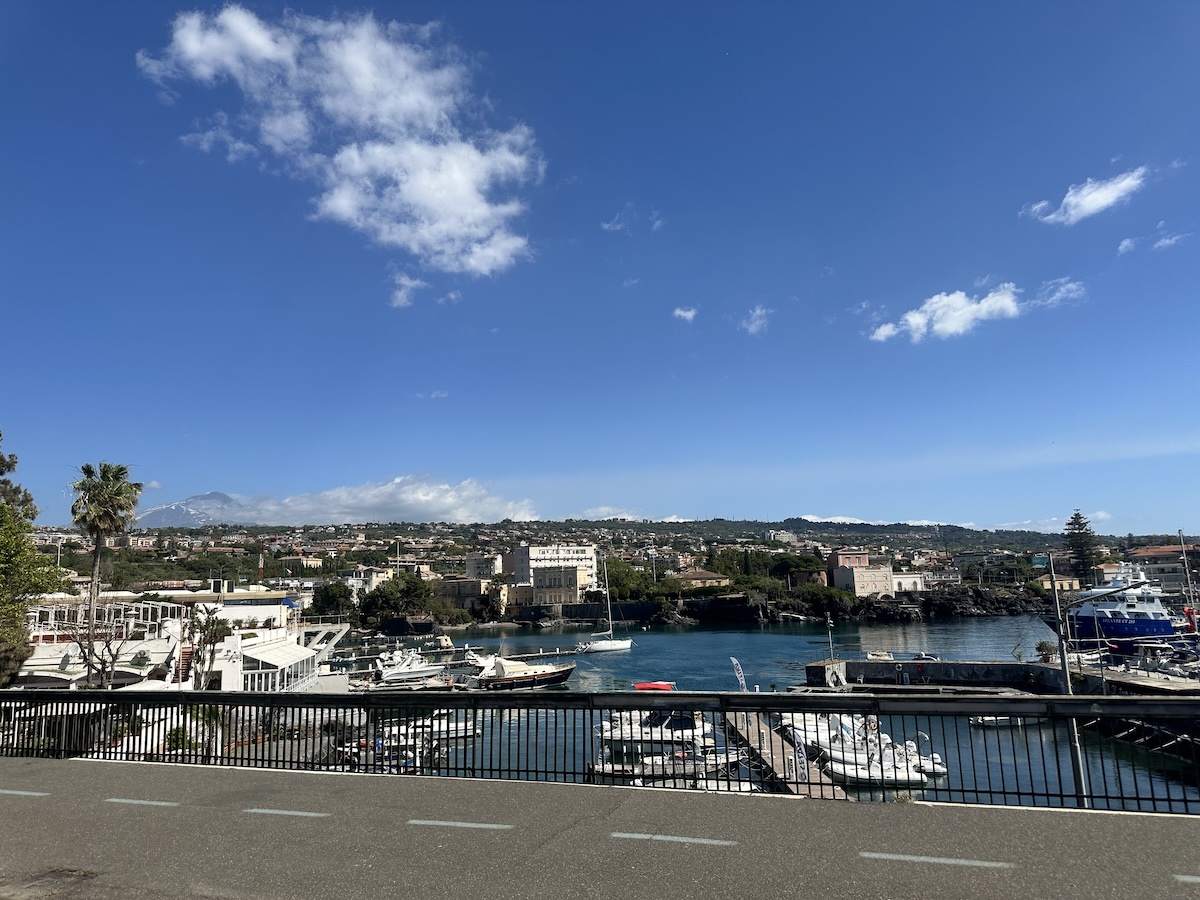 Blick auf den Catania-Hafen mit blauem Meer, weißen Wolken am Himmel und zahlreichen Booten an der Küste, im Hintergrund Gebäude und Vegetation.
