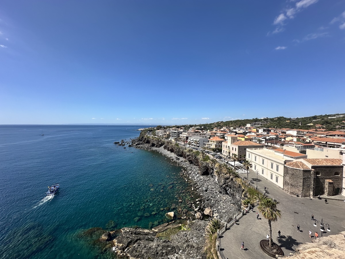 Türkisblaues Meer entlang der Küstenpromenade bei Catania, Blick auf Häuserdächer und Felsklippen unter klarem Himmel.