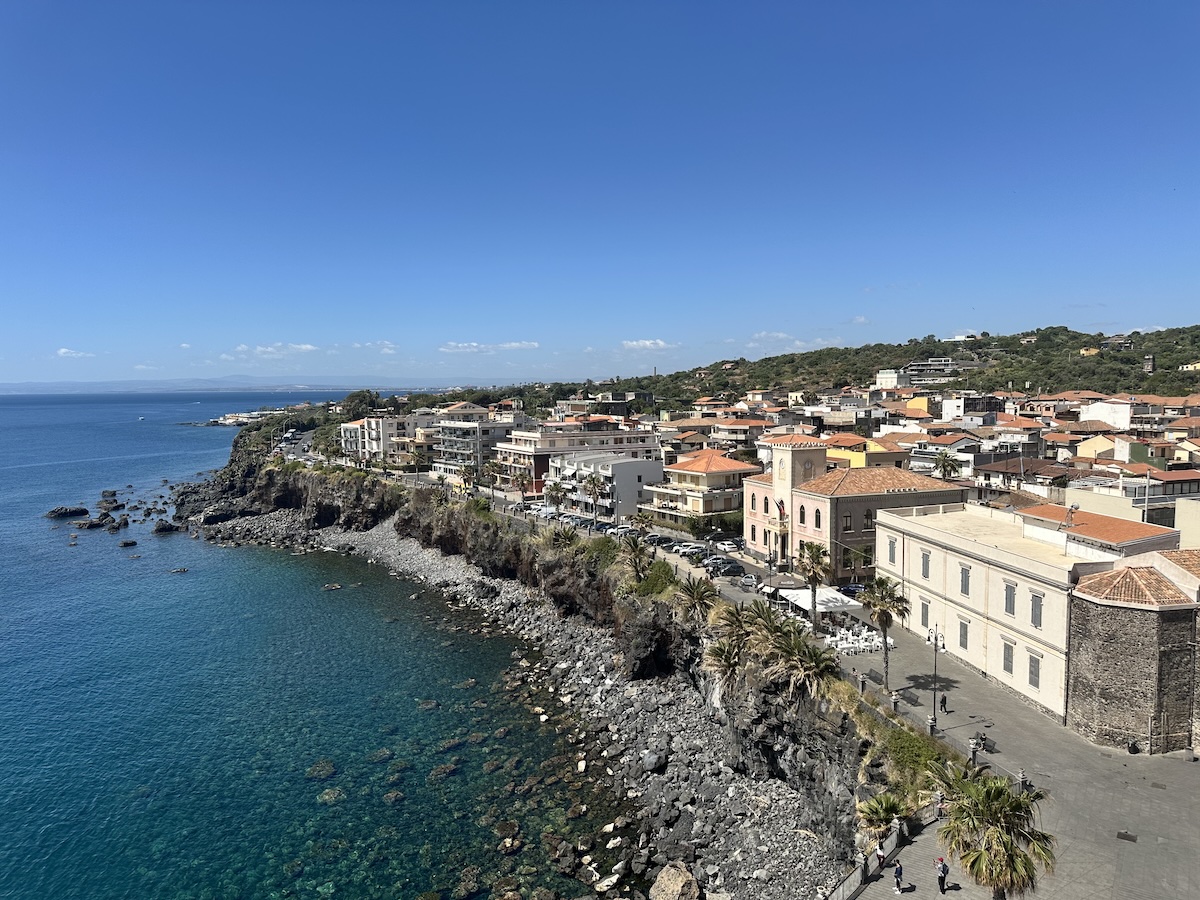 Blick auf Aci Castello: blaues Meer, schwarzer Lavastein an der Küste und Häuser mit roten Dächern unter klarem Himmel.