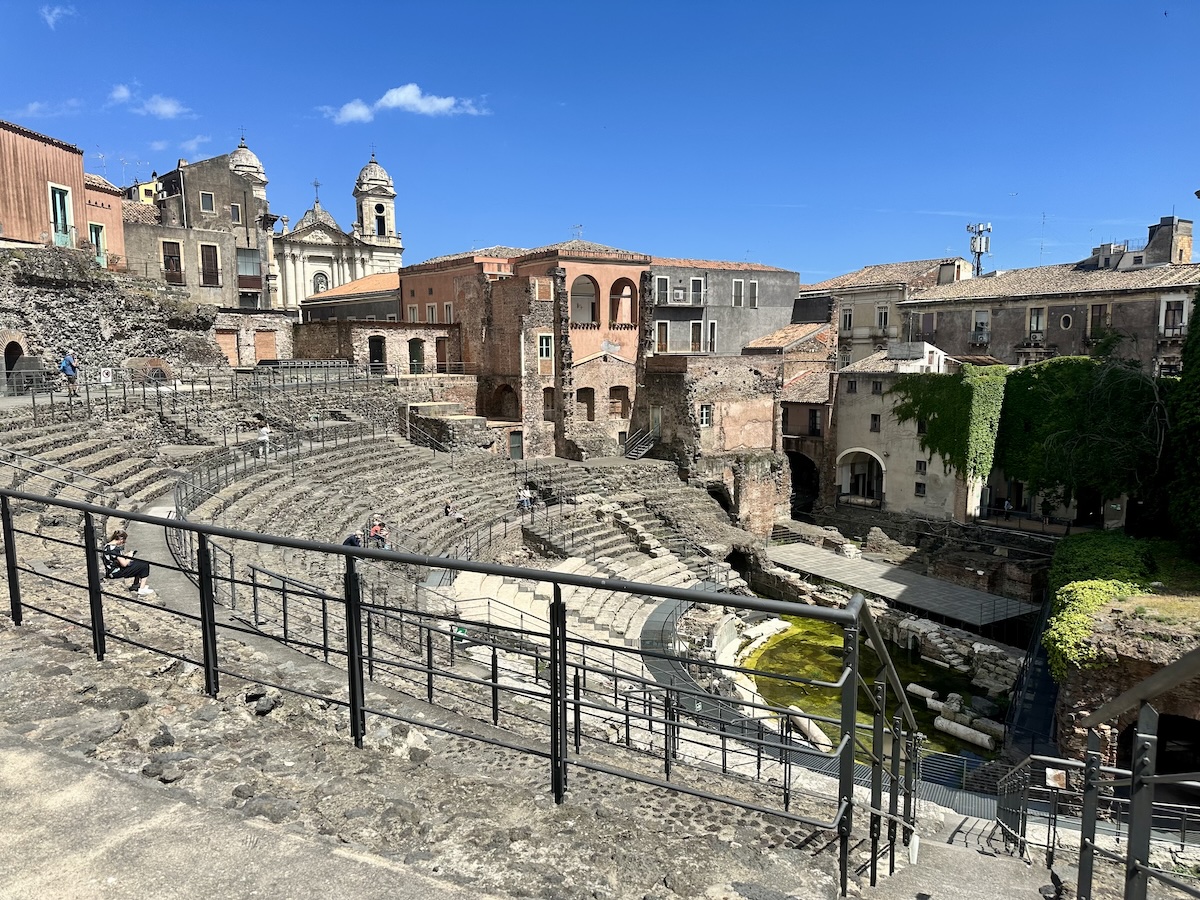 Steinerne Stufen und Lavasteinresten des Römischen Theaters von Catania unter blauem Himmel, mit historischer Altstadt und markanten Gebäuden im Hintergrund.