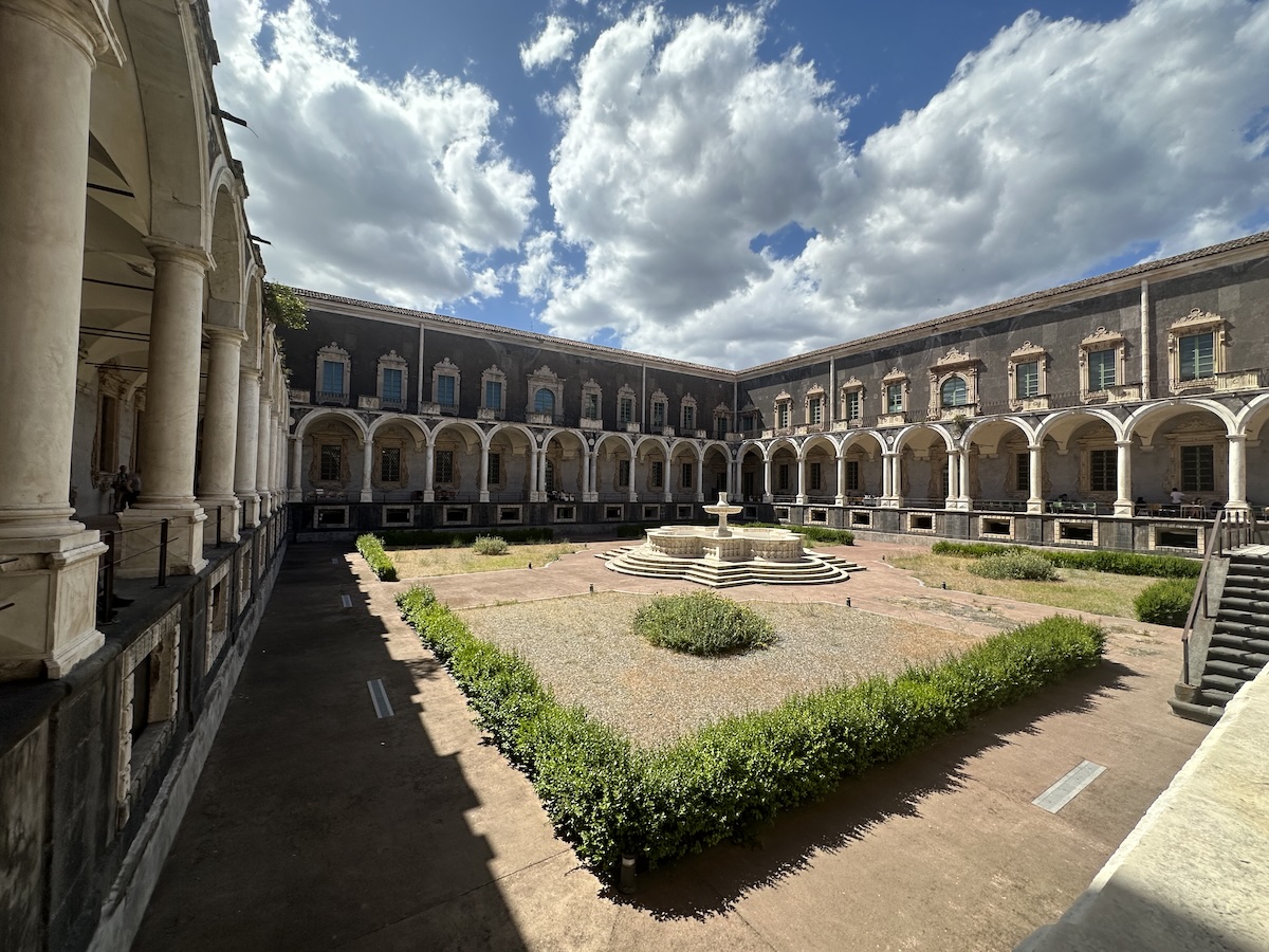 Weite Innenhofanlage mit gewölbten Arkaden im Kreuzgang des Benediktinerklosters San Nicolò l’Arena in Catania, hellgrauer Stein und Grünbeete.