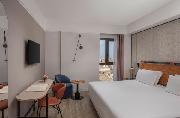 Helles, modern eingerichtetes Zimmer in Catania Centro mit weißem Bett, orangefarbenen Kissen und warmen Holzelementen, Blick zum Balkon und Vorhang.