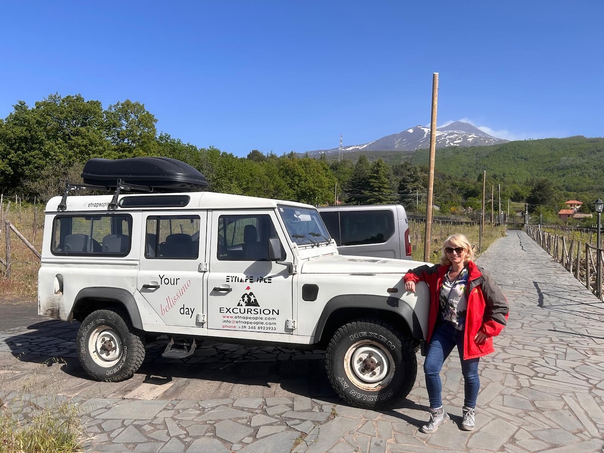 Vor einem schneebedeckten Ätna erhebt sich ein weißer Offroad-Jeep auf Steinweg, daneben steht eine Reisende in roter Jacke.