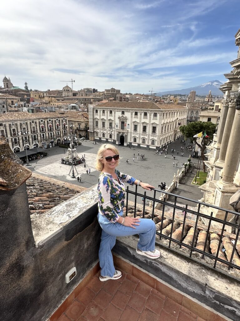 Mit Blick über die Piazza del Duomo in Catania sitzt eine Frau (Mandy) auf einer Dachterrasse, während der Ätna am Horizont aufragt.