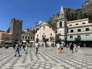 Auf der Piazza IX Aprile in Taormina stehen Besucher vor Kirche, Uhrturm und Palazzi unter blauem Himmel, während Dolce & Gabbana-Cafés die Altstadt säumen.