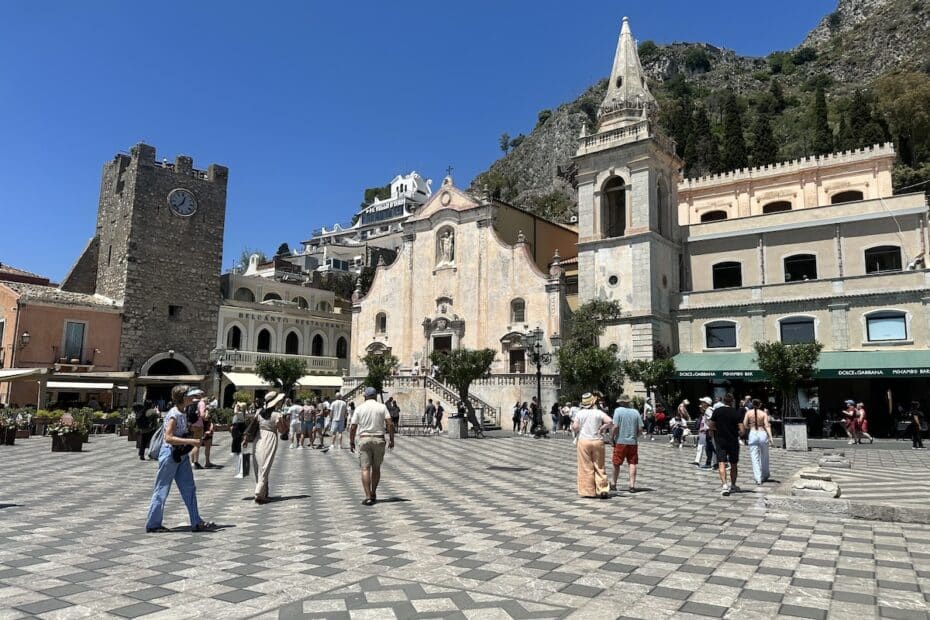 Auf der Piazza IX Aprile in Taormina stehen Besucher vor Kirche, Uhrturm und Palazzi unter blauem Himmel, während Dolce & Gabbana-Cafés die Altstadt säumen.