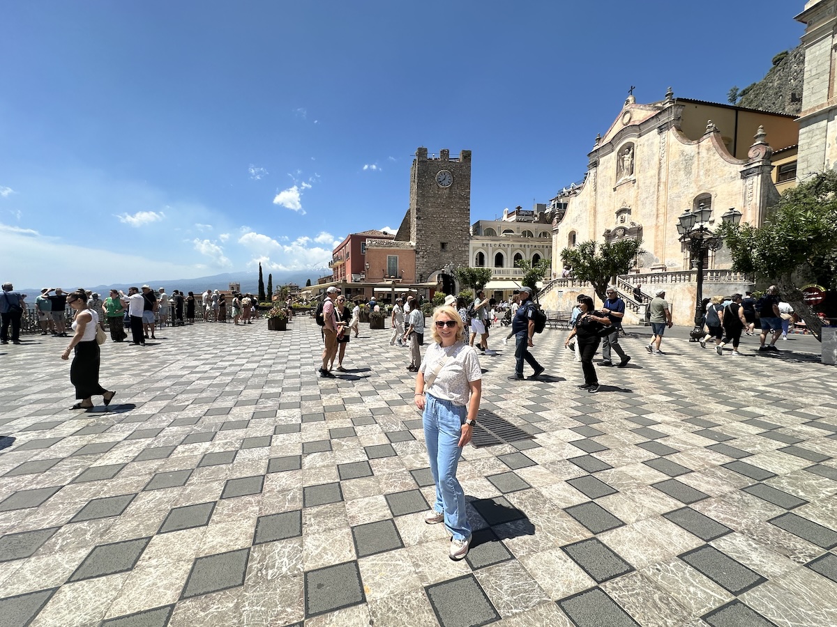 Am belebten Platz in Taormina stehen Reisende zwischen Uhrturm und Kirche, während der Blick über Sizilien bis zur Küste und zum Meer reicht.