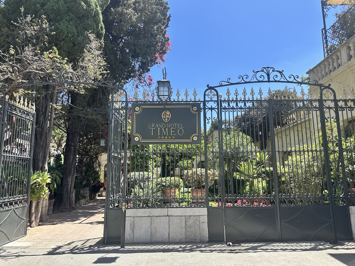 Vor dem Grand Hotel Timeo in Taormina führt ein schmiedeeisernes Tor zu einer grünen Gartenanlage unter klarem Himmel.