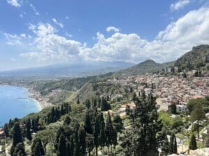 Über Taormina öffnet sich ein weiter Blick auf Altstadt, Küste und den Ätna am Horizont, eingerahmt von blauem Himmel und Wolken.