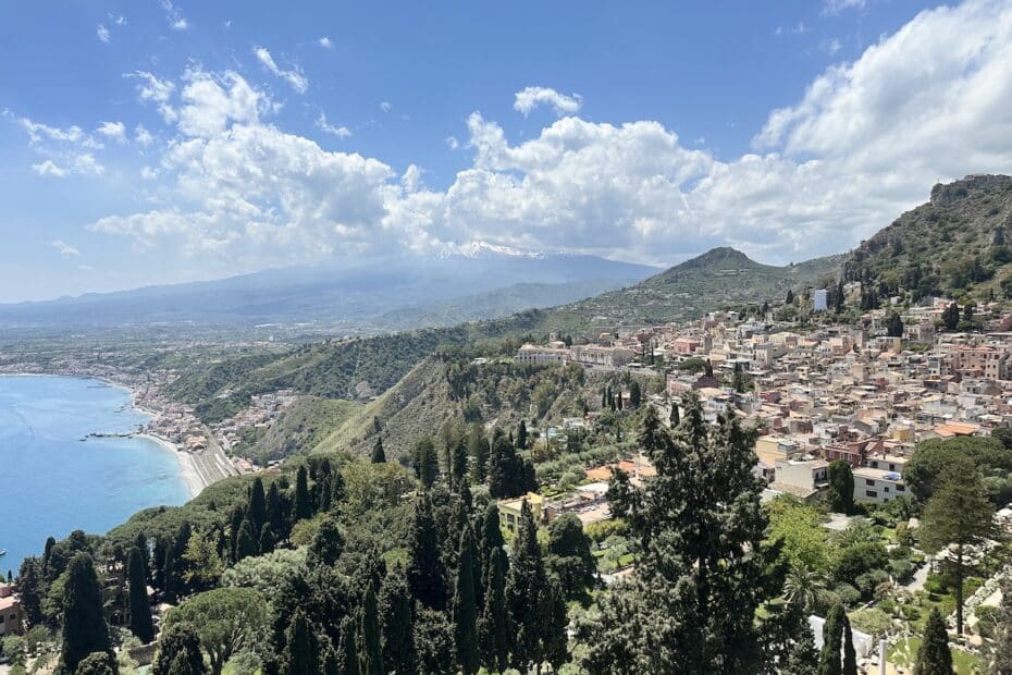 Über Taormina öffnet sich ein weiter Blick auf Altstadt, Küste und den Ätna am Horizont, eingerahmt von blauem Himmel und Wolken.