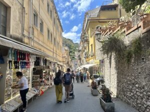 In Taorminas Altstadt schlendern Passanten an Souvenirläden, Cafés und alten Fassaden vorbei, während sich die enge Gasse zum Hügel hin öffnet.