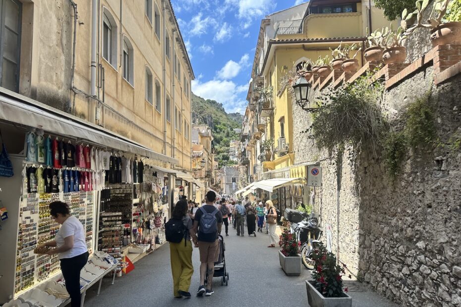 In Taorminas Altstadt schlendern Passanten an Souvenirläden, Cafés und alten Fassaden vorbei, während sich die enge Gasse zum Hügel hin öffnet.