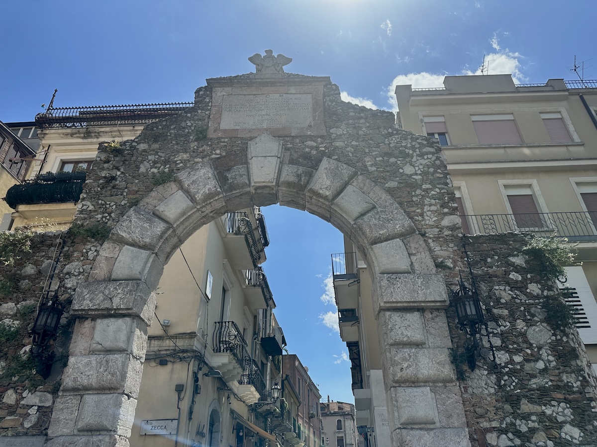 Unter dem steinernen Stadttor von Taormina führt der Blick in eine enge Altstadtgasse mit Häusern und blauem Himmel in Sizilien.
