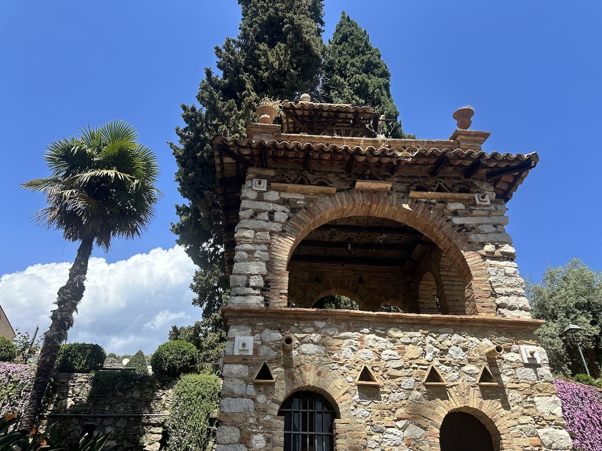 In Taormina steht ein historisches Steingebäude mit Rundbögen zwischen Palmen und Zypressen unter tiefblauem Himmel.