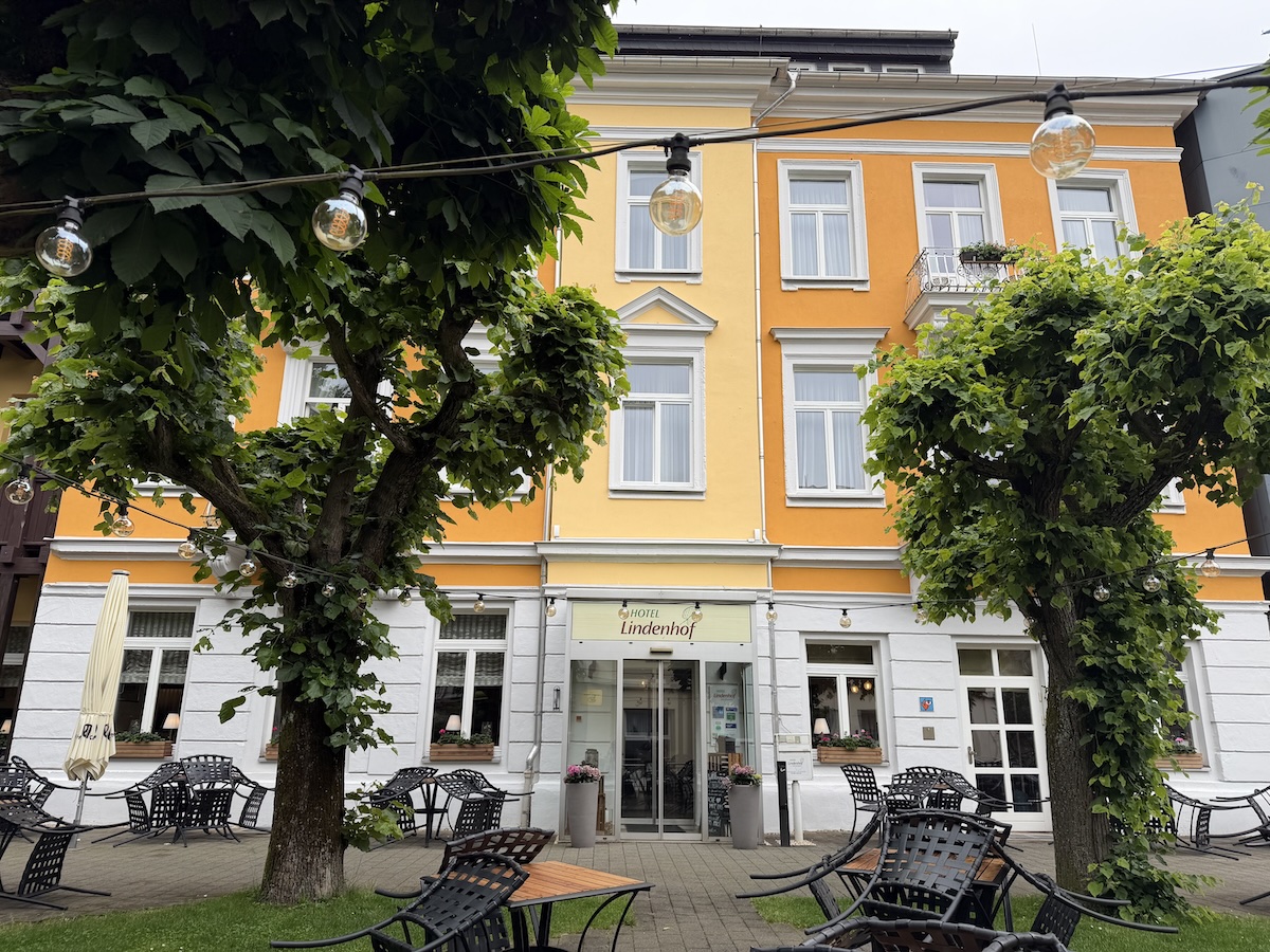 Im ruhigen Innenhof des Hotel Lindenhof in Bad Schandau stehen unter dichten Lindenbäumen Stühle, Tische und Lichterketten vor einer orangefarbenen Fassade.