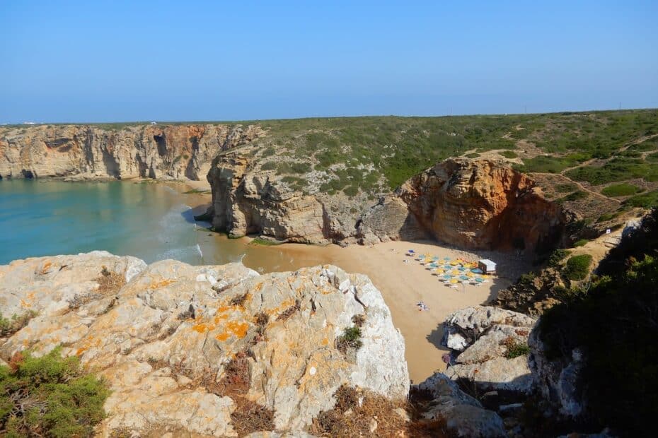 Unter hohen Kalkklippen liegt eine kleine Sandbucht mit klarem Wasser und bunten Sonnenschirmen an der Algarve.