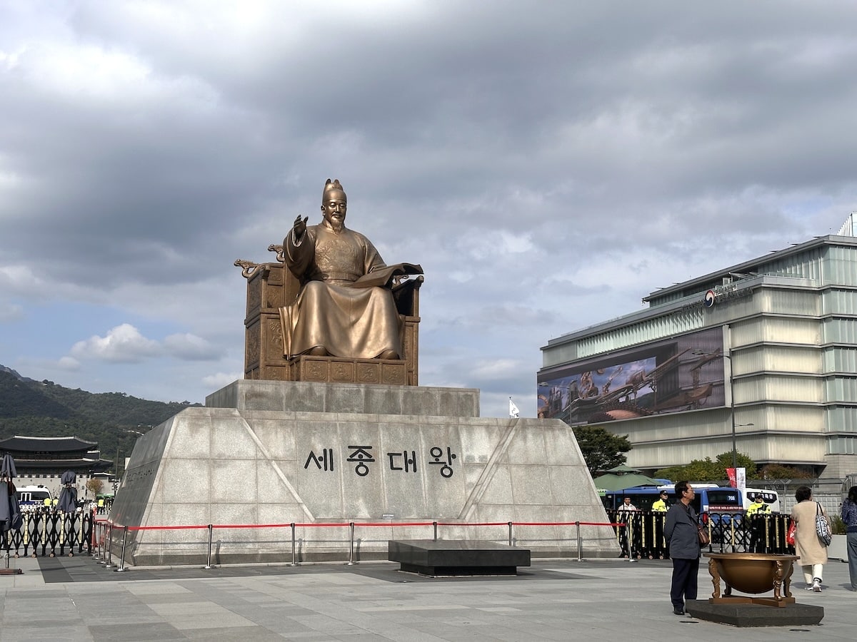 Die Bronze-Statue von Sejong dem Großen steht auf dem Gwanghwamun-Platz in Seoul vor dramatischem Himmel und moderner Architektur.