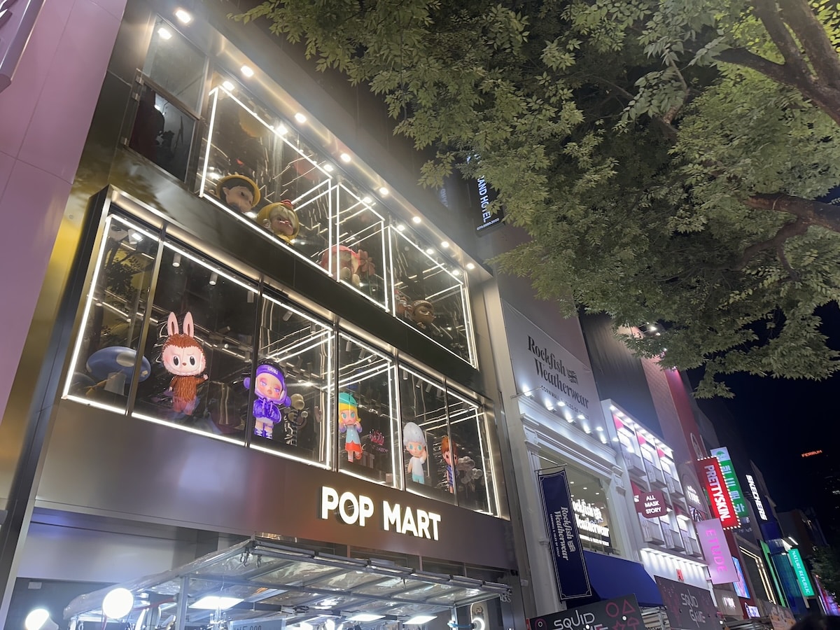 Das leuchtende POP MART-Schaufenster bei Nacht zeigt Figuren hinter Glas, daneben weitere Ladenfronten und ein dichter Baum über der Straße.