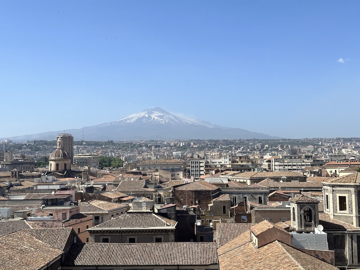 Weitblick über die Dächer von Catania mit einer Kirchenkuppel im Vordergrund und dem schneebedeckten Ätna unter klarem Himmel im Hintergrund.