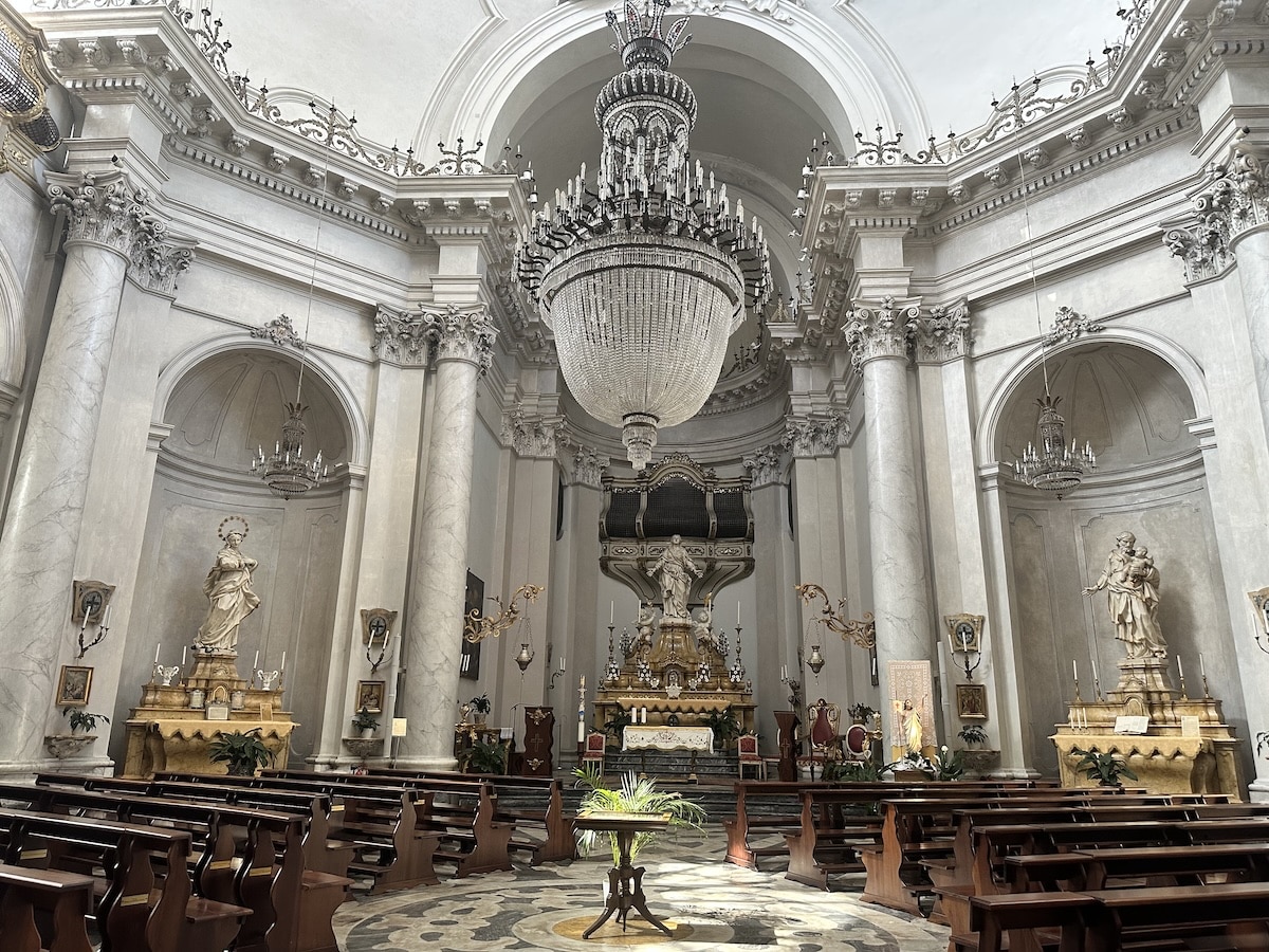 Der helle Kirchenraum zeigt einen prachtvollen barocken Altar mit Marmorsäulen, großen Kronleuchtern und langen Holzbänken vor dem Mittelgang.