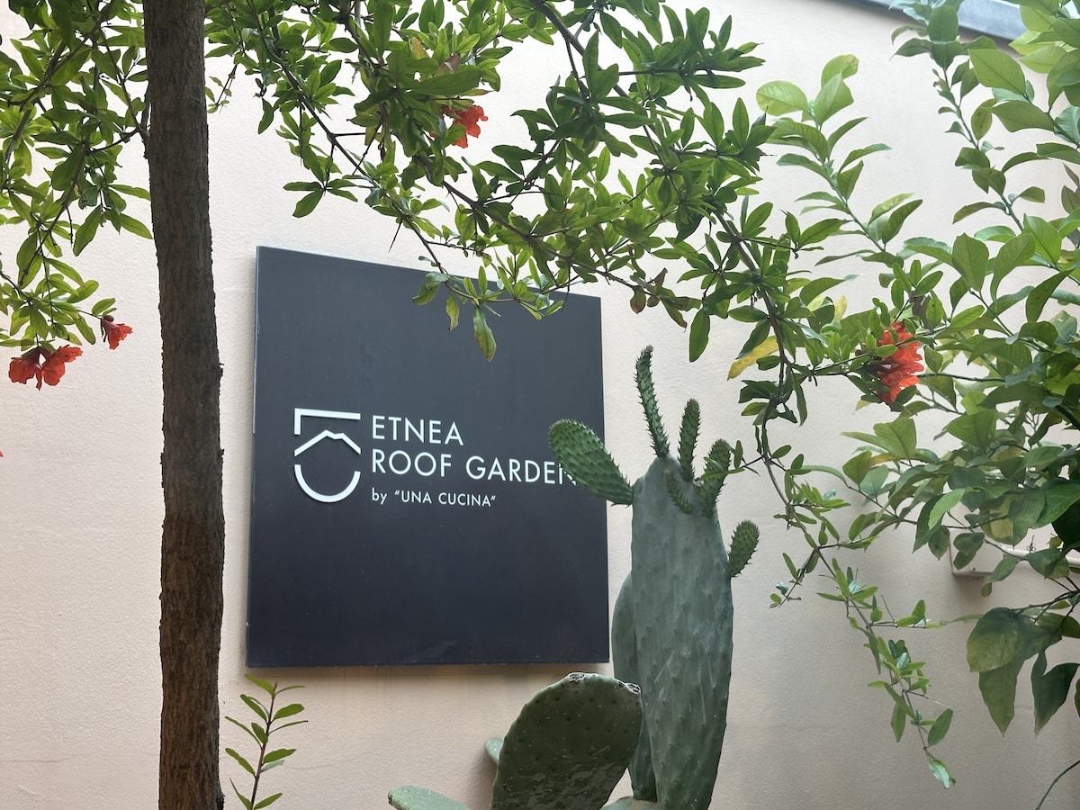Vor einer hellen Wand hängt ein schwarzes Schild mit der Aufschrift "ETNEA ROOF GARDEN by UNA CUCINA", umrahmt von grünen Pflanzen und roten Blüten.