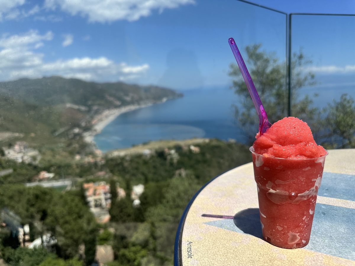 Vor dem Blick auf die Bucht von Taormina steht ein roter Granita-Becher mit violettem Löffel auf einem Tisch.