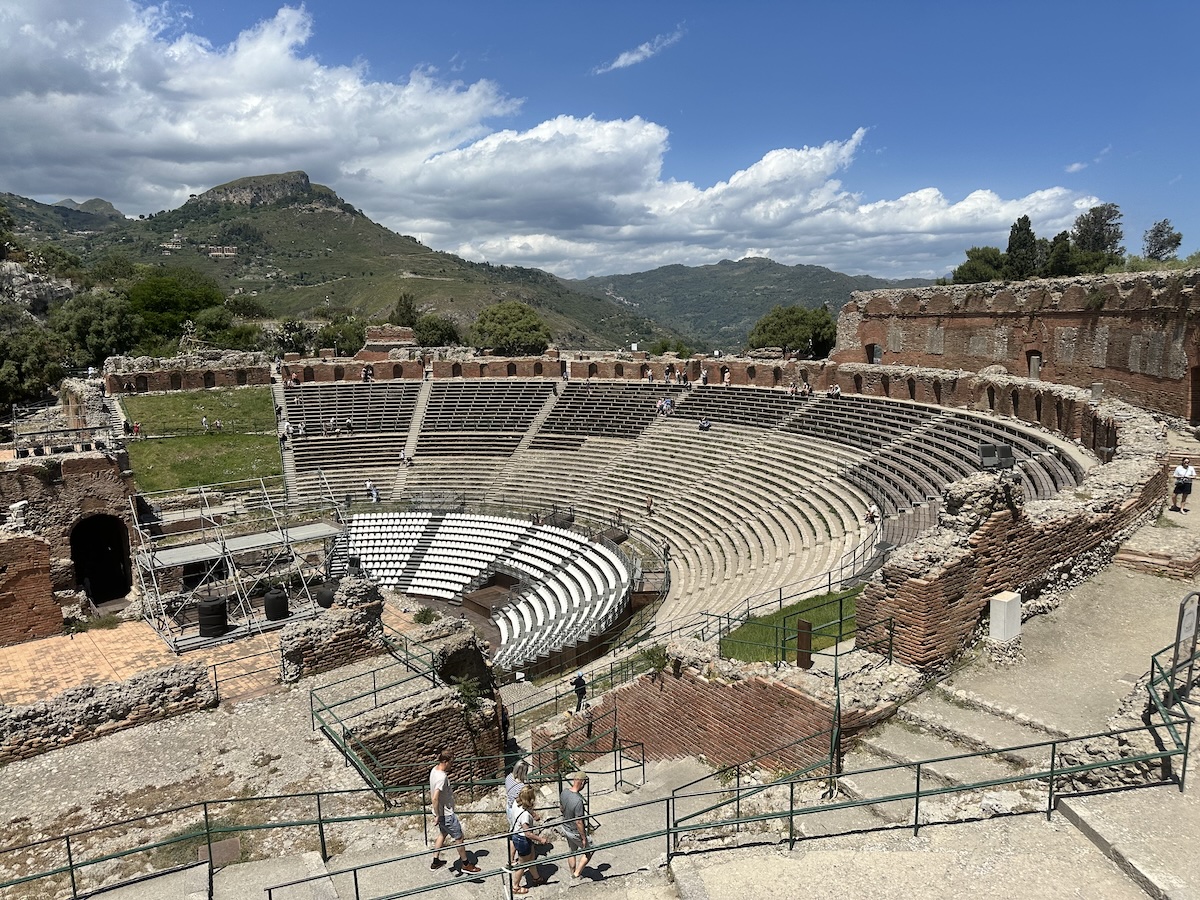 Im Teatro Antico Taormina liegen die steinernen Zuschauerreihen offen vor einer grünen Berglandschaft unter blauem Himmel.