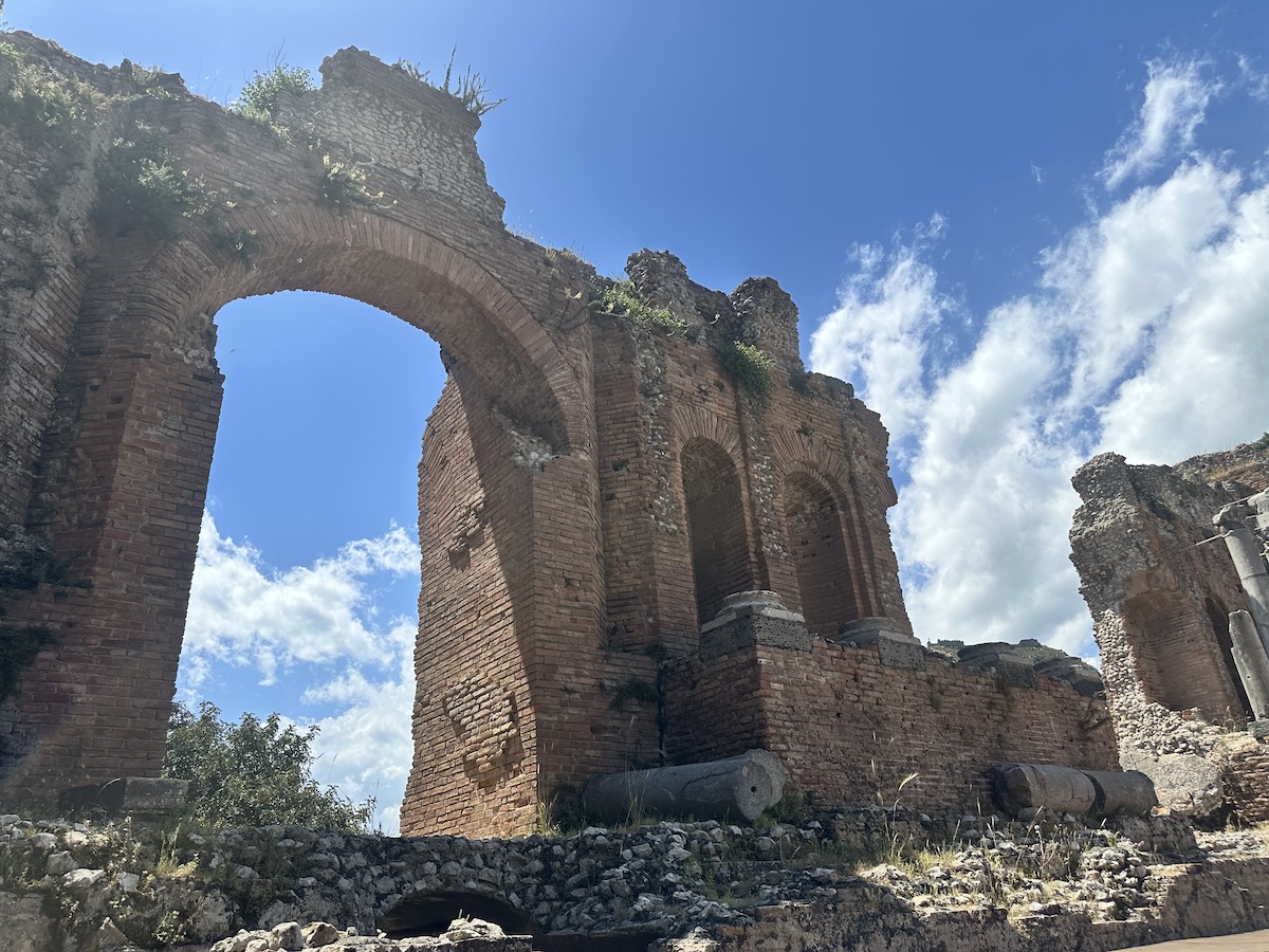 Im Teatro Antico Taormina erhebt sich ein großer Backsteinbogen zwischen antiken Mauern unter blauem Himmel.