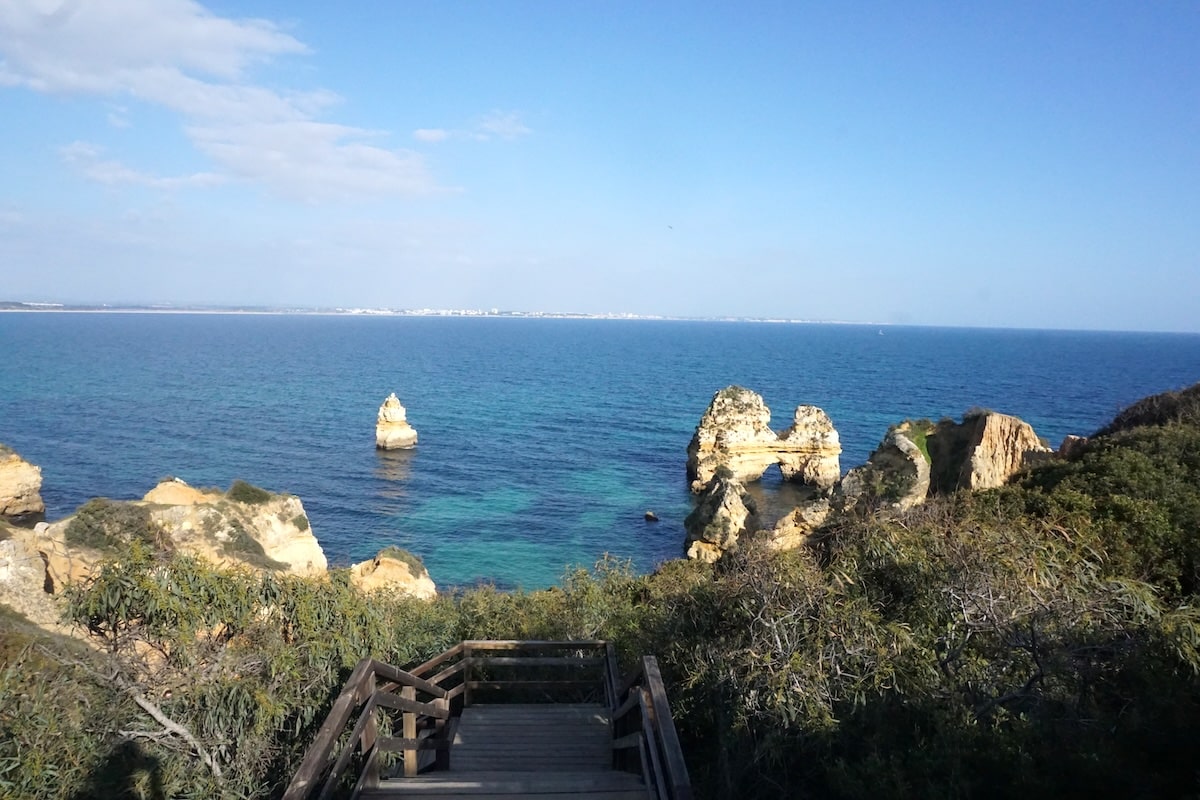 An der Küste von Lagos fällt der Blick auf türkisblaues Wasser, markante Felsen und einen hölzernen Treppenabgang. Praia do Camilo
