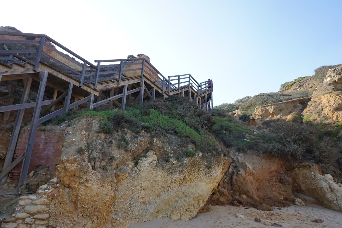 Ein hölzerner Steg führt über die Felsküste von Lagos; unten öffnet sich ein kleiner Sandstrand zwischen den Klippen.
