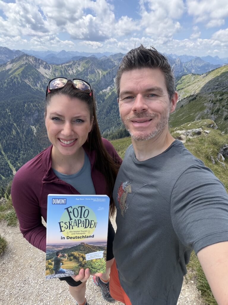 Biggi und Florian von Phototravellers zeigen vor alpiner Berglandschaft ihr Buch „Foto-Eskapaden in Deutschland“.